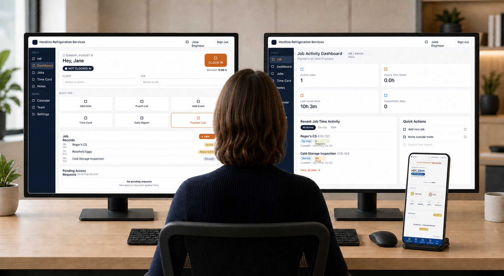 Office manager reviewing the TradeLog Site project activity dashboard on two monitors with the mobile app open beside them.